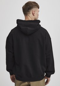 Sudadera negra de gran tamaño con una capucha grande y puños acanalados. Confeccionada con un material suave de textura lisa y sin patrones visibles.
