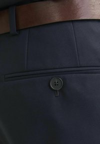 Tumesinine kangast püksid, millel on sile tekstuur, koos õmmeldaud tasku ja musta nupuga detailiga. Komplektis on pruuni nahast vöö.