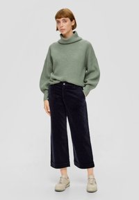 Groene ribgebreide trui met hoge kraag, gecombineerd met donkerblauwe wijde corduroypantalon en lichtbeige sneakers.