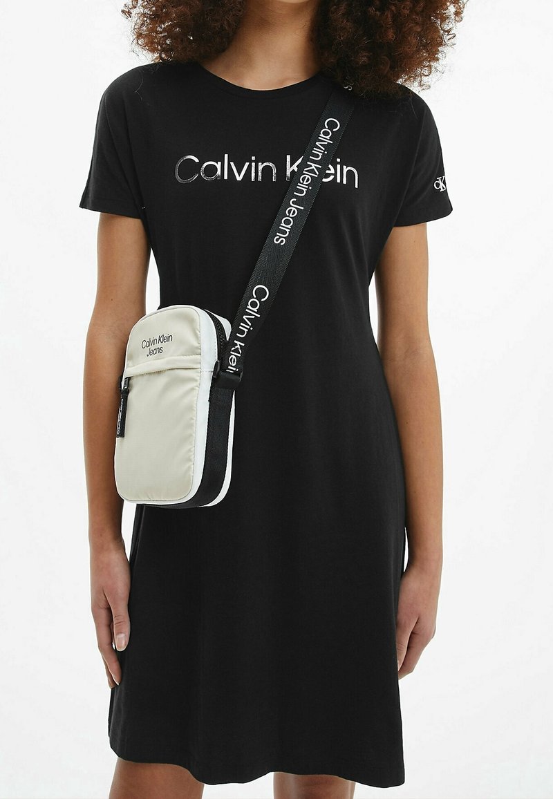Person, die ein schwarzes Calvin Klein T-Shirt-Kleid und eine beige Calvin Klein Umhängetasche, die quer über die Brust hängt, trägt.