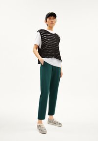 Zwarte gebreide mouwloze top over een wit T-shirt, gecombineerd met groene tapered broek en zwart-wit gestreepte sneakers.