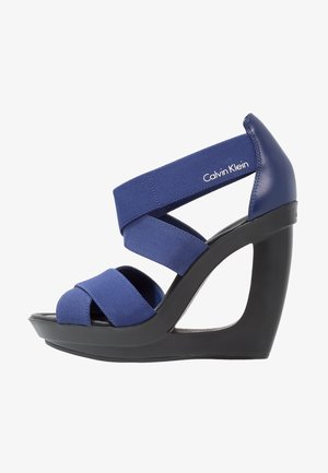 Sandales à talons compensés bleu marine avec des sangles élastiques, design à bout ouvert, et une plateforme noire contrastante. Elles présentent un talon courbé et un détail de logo.