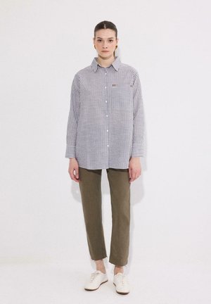 Η γυναίκα στέκεται φορώντας μακρυμάνικο πουκάμισο με κάθετες ρίγες και κουμπιά, ελαφρώς ίσια παντελόνια σε ελαιί πράσινο, λευκά αθλητικά παπούτσια, με τα μαλλιά μαζεμένα πίσω και χρυσούς κρίκους στα αυτιά.