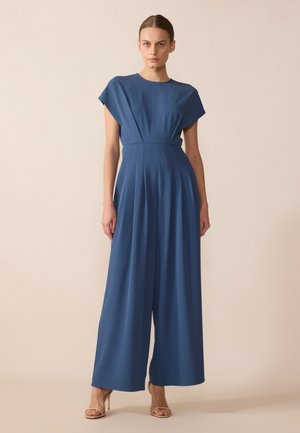 Blå, bredbenet jumpsuit med korte ærmer og en tætsiddende talje, der har plisseringer og en glat tekstur. Båret med lyse hæl sandaler.