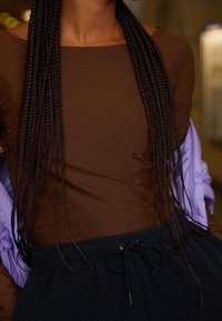 Detailní záběr na osobu oblečenou v hnědém dlouhém rukávu s jemnými vzory, černých kalhotách na šňůrku a světle fialové bundě přehozené přes ramena.