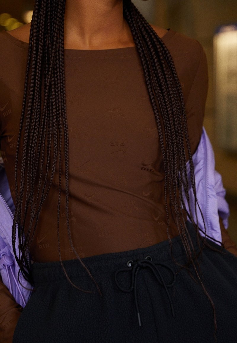 Detailní záběr na osobu oblečenou v hnědém dlouhém rukávu s jemnými vzory, černých kalhotách na šňůrku a světle fialové bundě přehozené přes ramena.