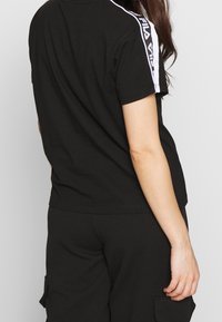 Schwarzes T-Shirt mit kurzen Ärmeln und Rundhalsausschnitt, mit weißen Logo-Akzenten an den Schultern. Der Stoff wirkt glatt und lässig.