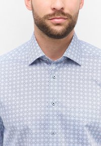 Uomo con barba che indossa una camicia azzurra chiara con motivo floreale circolare blu scuro, mostrato dalla collana fino a metà torace.