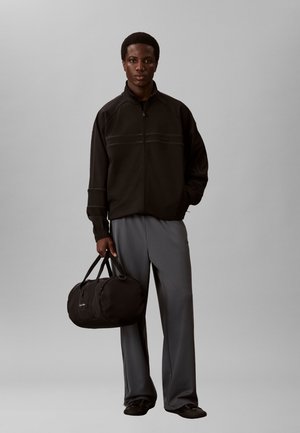 Nuori mies seisoo musta vetoketjutakki päällä, harmaat leveälahkeiset housut ja mustat kengät jalassa, pitäen yhdellä kädellä mustaa duffel-laukkua.