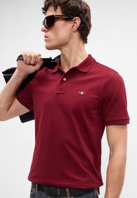 Burgundské polo tričko s klasickým golierom, krátkymi rukávmi a dvoma gombíkmi. Obsahuje malé logo na hrudi a hladkú textúru.