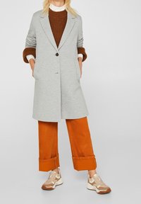 Mujer vistiendo un abrigo gris de un solo botón sobre un suéter marrón, pantalones de pierna ancha naranjas recogidos y zapatillas beige, de pie con las manos en los bolsillos.