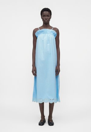 Γυναίκα φοράει αμάνικο, ανοιχτό μπλε midi φόρεμα με λεπτομέρεια βολάν και τελείωμα με τρυπητό σχέδιο, σε συνδυασμό με μαύρα παπούτσια Mary Jane, στέκεται μπροστά σε λευκό φόντο.