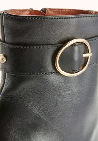 Bottines en cuir noir avec une forme courbée, accent en métal doré de forme circulaire et couture visible autour du col.