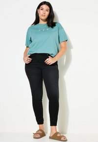 Camiseta de algodón color teal con mangas cortas y diseño bordado de hoja de palma, combinada con vaqueros ajustados negros de tiro alto y sandalias marrones tipo slide.