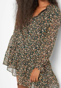 Svart blommig klänning med långa, genomskinliga ärmar. Har en volangkrage och en svängande nederdel prydd med små orange, vita och gröna blommor.