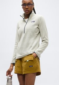 Γυναίκα που φοράει μπεζ fleece με σχέδιο και φερμουάρ στο ένα τέταρτο, μουσταρδί cargo σορτς, γυαλιά ηλίου, κρατώντας ένα ανοιχτό γκρι παγούρι, σε απλό φόντο.