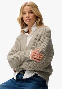 Blondynka w grubym beżowym swetrze wzoru dzianiny, nosząca białą koszulę z guzikami i niebieskie dżinsy, z założonymi na siebie ramionami, patrzy w bok.
