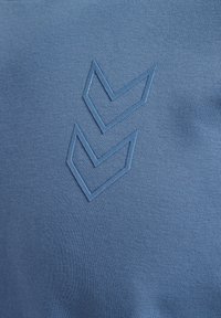 Twee marineblauwe chevrons geperst op zacht blauw gebreid stof, waarbij de textuur van de stof en subtiele schaduwen zichtbaar zijn.