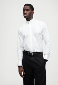 Hvit langermet skjorte med ståkrage, sammen med svarte bukser og et svart belte, med sølvfargede detaljer og hvit søm.