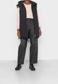 Czarna bezrękawnikowa kamizelka puchowa z kapturem, przednim zamkiem błyskawicznym i detalami ze sznurkiem, noszona na różowej w paski bluzce z długim rękawem oraz ciemnych, szerokich dżinsach.