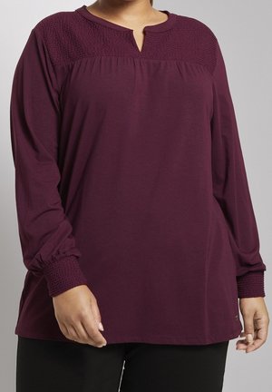 Femme portant une tunique prune à manches longues avec un empiècement texturé et des poignets, debout devant un fond neutre.