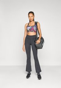Stanik sportowy w kolorowy wzór motyli, czarne legginsy z wysokim stanem i szerokimi nogawkami oraz czarne buty sportowe. Modelka trzyma ciemną torbę na sprzęt.