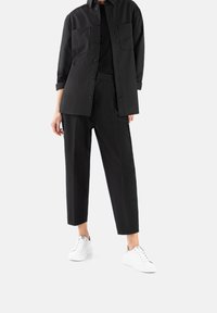 Black shirt and trousers made of smooth fabric, featuring a button-up design, two chest pockets, and white sneakers for contrast.