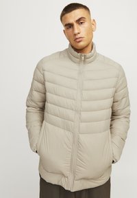 Beige puffjakke med høj krave, vertikalt kviltet design og lynlås-lukning. Har sidelommer og en glat, blank overflade.