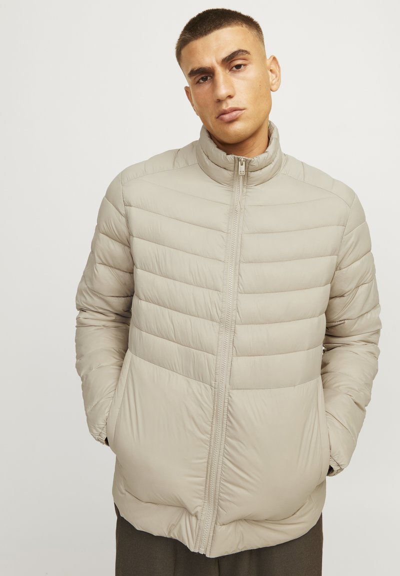 Beige puffjakke med høj krave, vertikalt kviltet design og lynlås-lukning. Har sidelommer og en glat, blank overflade.