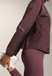 Bordeaux lichte jas met hoge kraag, elastische manchetten en reflecterende accenten, gecombineerd met bijpassende leggings en witte sneakers.