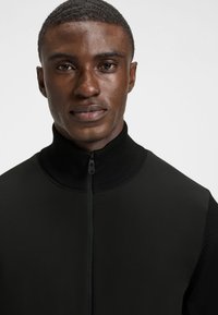 Juodas liemenė su užtrauktuku ir aukšta apykakle. Pagaminta iš lygaus audinio su tekstūruota megzta apykakle. Turi centrinį užtrauktuką ir minimalistinį dizainą.