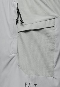 Ljusgrå jacka med en texturerad ficka i rutmönster. Inkluderar svarta dragsnören och en tryckt logotyp längst ner. Slät tygfinish.