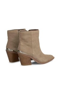 Botas de tobillo de ante beige con punta afilada, con un acento metálico en la parte trasera y un tacón de madera apilada. Cierre de cremallera en el costado.
