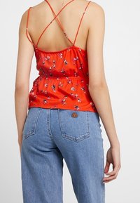 Rode bloemenprint cami-top met verstelbare gekruiste bandjes en een gesmokte taille. Gecombineerd met lichtblauwe high-waisted jeans.