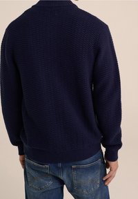 Pull tricoté bleu marine avec un tissu texturé, col, poignets et ourlet côtelés. Porté avec un jean bleu clair agrémenté d'accents sur les poches arrière.