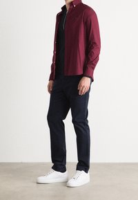 Bordowa koszula zapinana na guziki z kołnierzykiem, zestawiona z czarną koszulą. Ciemne spodnie i białe sneakersy dopełniają strój. Prosty, nowoczesny design.