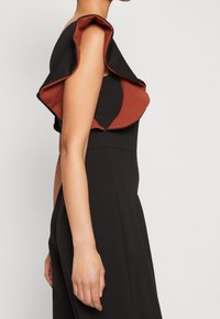 Mujer vestida con un vestido negro sin mangas con un detalle de volantes en capas de color óxido en el hombro y la parte superior del brazo, mostrada de perfil contra un fondo liso.