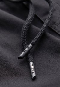 Gros plan sur des lacets de capuche tressés noirs avec des embouts métalliques gravés de « ondre » sur tissu noir.