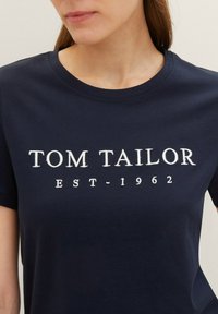 T-shirt en coton bleu marine foncé avec col rond et manches courtes. Présente un texte imprimé en blanc : "TOM TAILOR EST-1962." Texture lisse.
