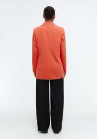 Blazer de coral con un corte ajustado y solapas de muesca, combinado con pantalones negros de pierna ancha. Textura de tela suave, diseño moderno.