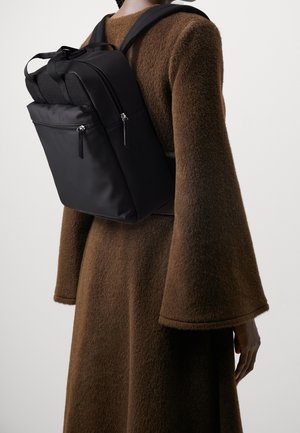 Persoon met een bruine, gestructureerde jas met brede mouwen en een zwarte, rechthoekige rugzak met ritsvakken op de rug.