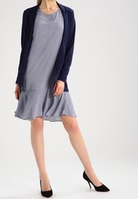 Námornická sako přes světle modrou šaty s puntíky a rozšířeným lemem. Černé vysoké podpatky dokončují outfit. Hladká textura po celé délce.