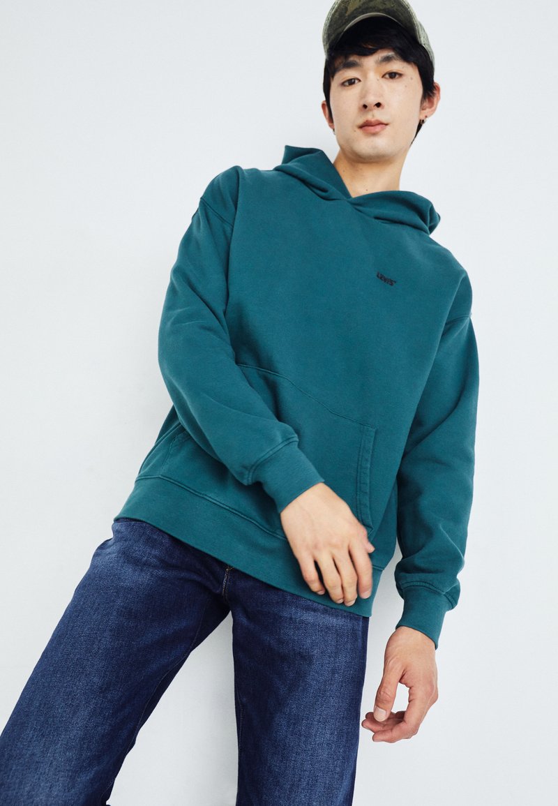 Sudadera verde azulado hecha de una tela suave, con un bolsillo frontal y capucha ajustable, combinada con jeans de mezclilla azules.