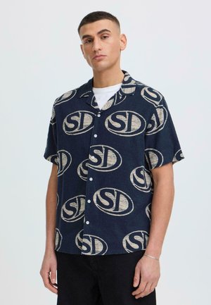 Młody mężczyzna w granatowej koszuli z guzikami, ozdobionej beżowymi logo „SD”, stojący na jednolitym jasnym tle.