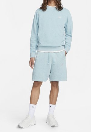 Person trägt ein hellblaues Nike-Sweatshirt und passende Shorts, weiße Nike-Socken und weiße Sneaker, steht mit einer Hand in der Tasche.