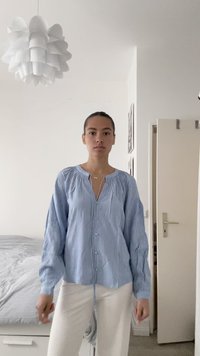 Sötét hajú nő, aki hátul összefogta a haját, kék csíkos gombos blúzt és fehér nadrágot visel, egy minimális kialakítású hálószobában áll, ahol egy ágy és egy lámpaernyő található.