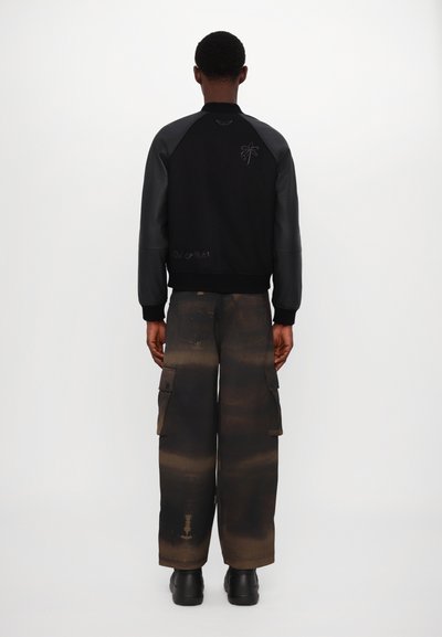 Veste bomber noire avec des manches en cuir, ornée d'une broderie de palmier. Pantalons cargo avec un motif dégradé beige et marron foncé. Chaussures noires.