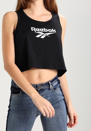 Kobieta ubrana w czarny, krótki top Reebok i niebieskie dżinsy z wysokim stanem oraz widocznym pępkiem na jednolitym tle.