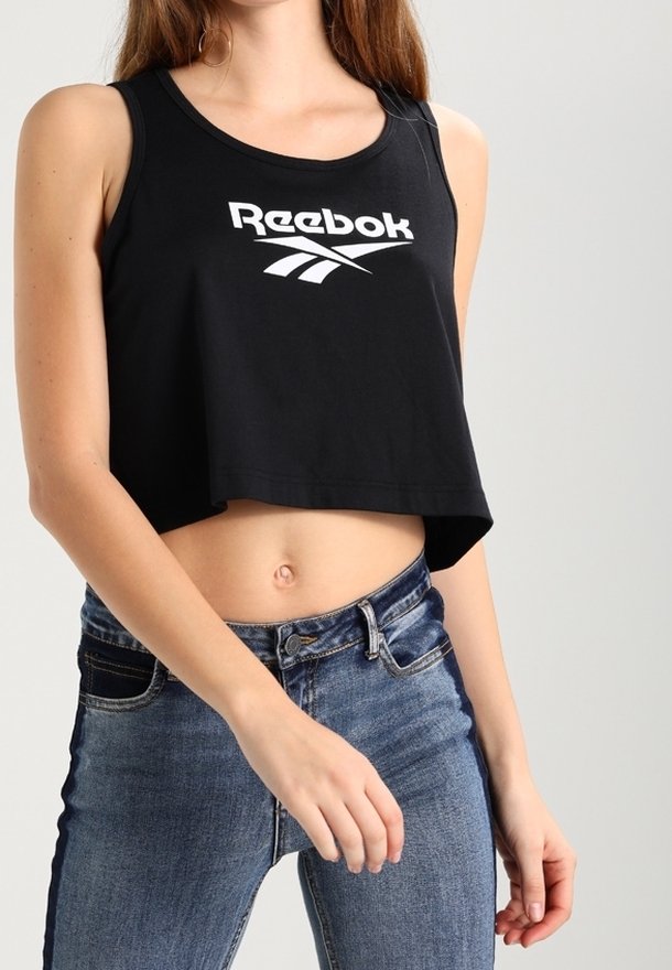 Femme portant un débardeur Reebok noir court et un jean bleu taille haute avec le nombril visible, sur fond uni.