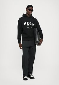 Felpa nera con logo bianco "MSGM MILANO", abbinata a pantaloni scuri larghi e scarpe nere con accenti bianchi. Modello che tiene una borsa nera.